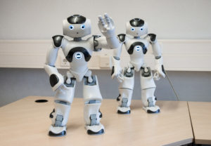 Picture of two NAO Robots. The robot on the left is waving.