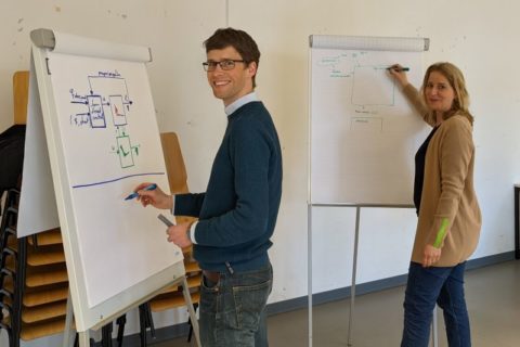 Picture of two people drawing models on flip charts and looking into the camera.