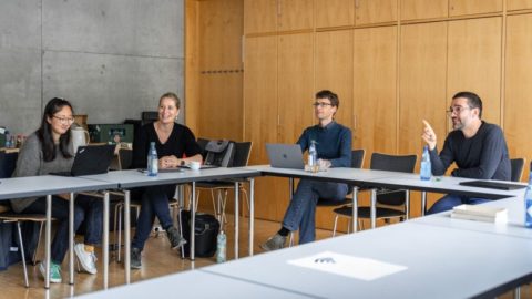 4 people are sitting at a table looking to the left, discussing and point to something outside of the frame