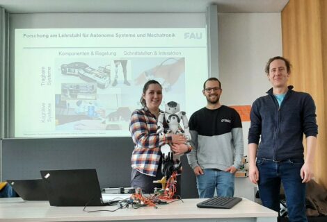 Picture with three people from the chair of autonomous systems and mechatronics. The person on the left is holding a humanoid robot. On the table in front of them is a robotic hand